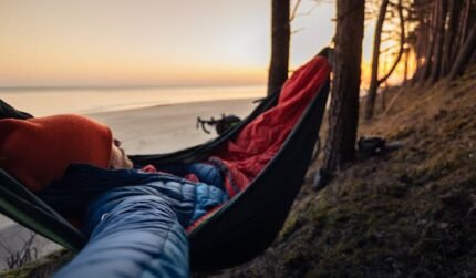 Person sleeping in hammock on forested beach at sunset, enjoying outdoor leisure.
